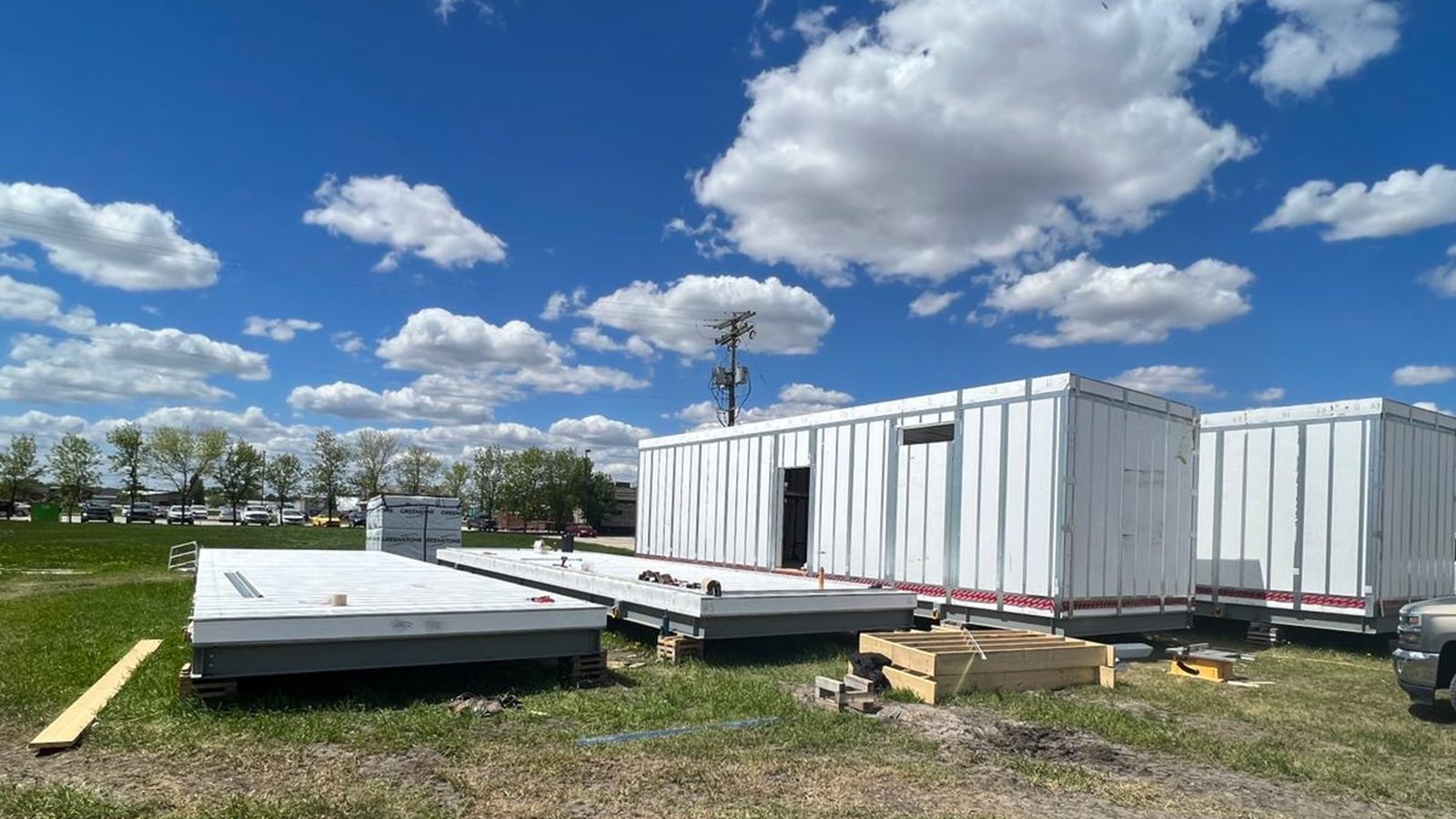 Modular home sections being assembled on a grassy lot under a bright blue sky, with wall modules and floor panels in place, a wheel loader, and a pickup truck