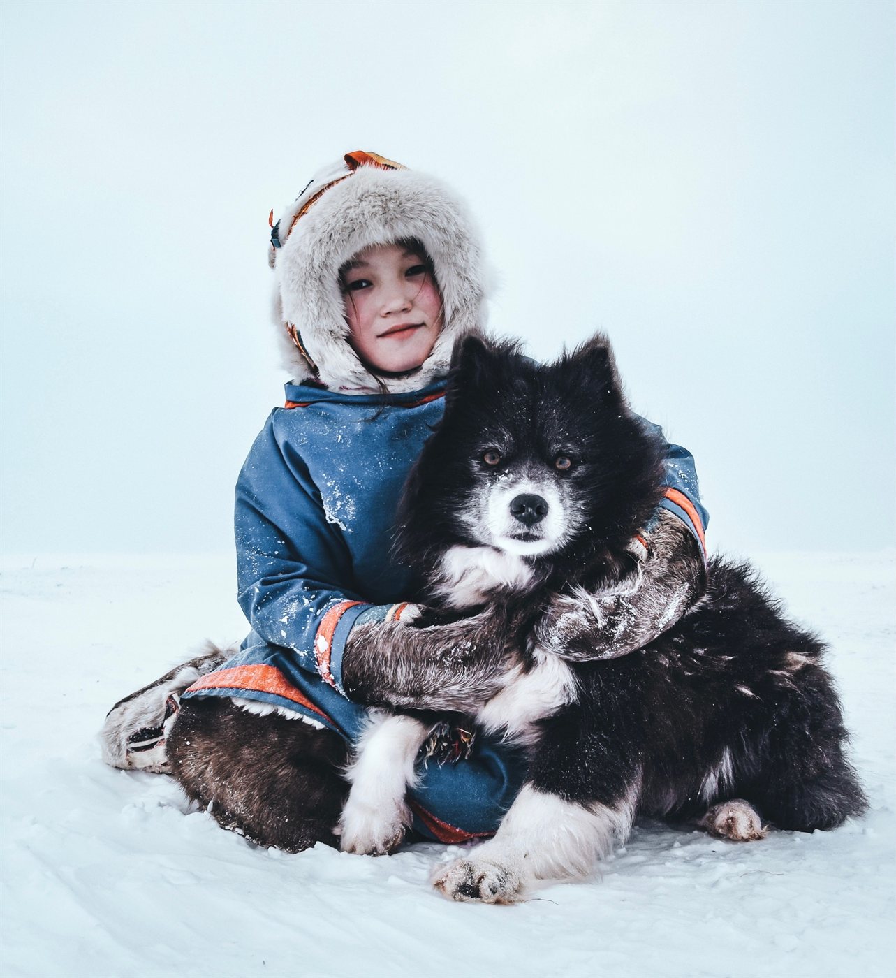 A young Indigenous child in a blue parka and thick fur-trimmed hood sitting in the snow, arms wrapped around a black and white sled dog resting against them