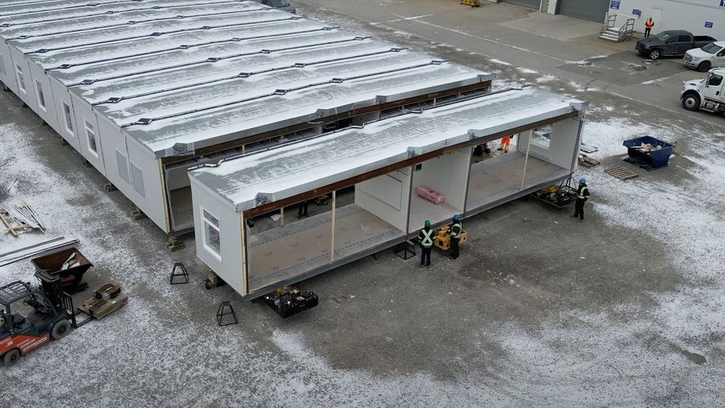 Modular building units being assembled in a snow-covered factory yard, with construction crews in high-visibility gear, a forklift, and delivery trucks