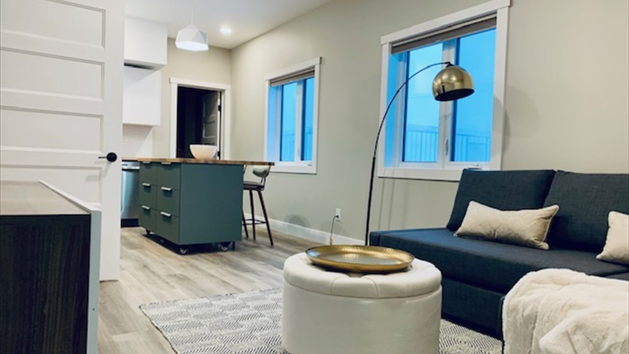 The interior of a finished modular home, a bright open living room with a dark sofa and cushions, a round ottoman on a patterned rug, and a kitchen island with an eating bar and a window behind