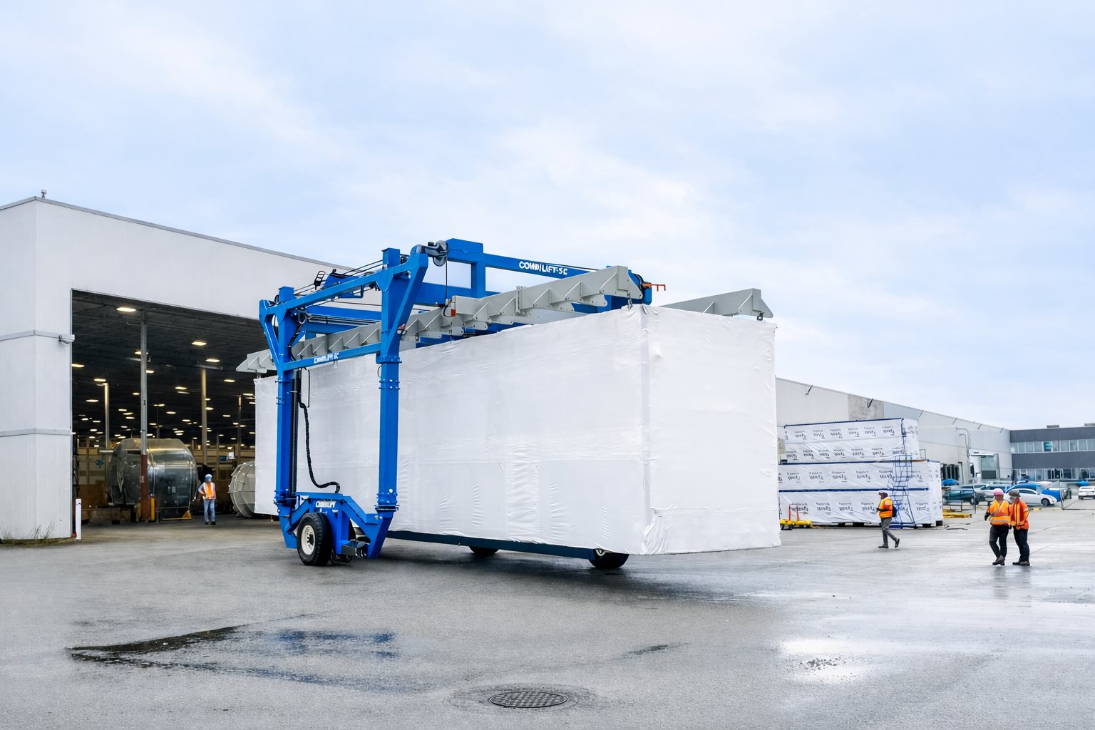 A large shrink-wrapped modular building unit being carried out of a factory by a blue straddle carrier, with crews in high-visibility gear