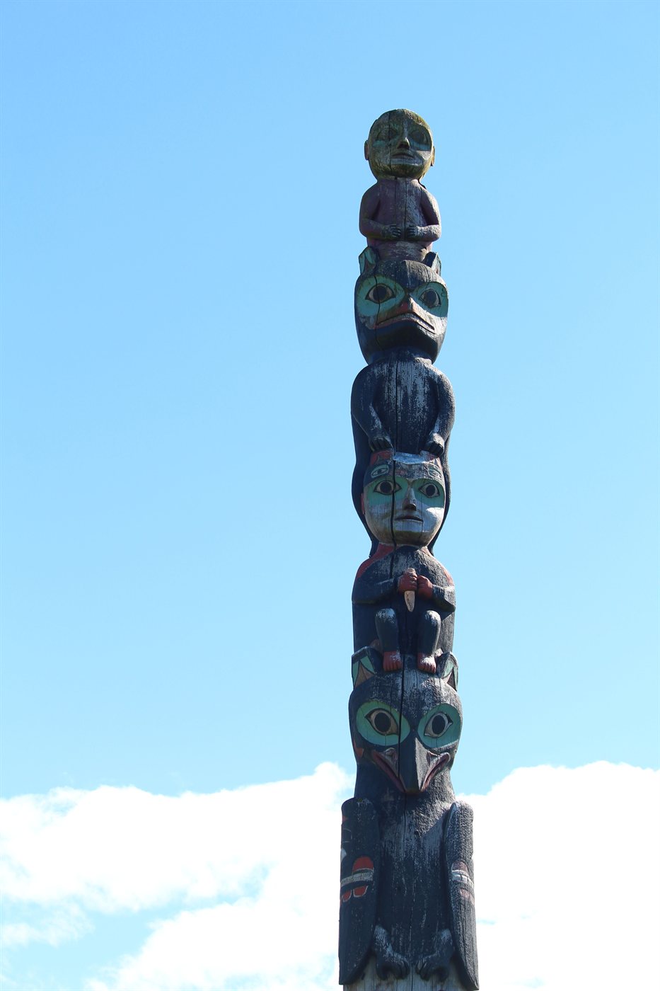 A Northwest Coast totem pole against a bright blue sky, carved with stacked figures including a bear, a human face with formline eyes, and a thunderbird, painted in teal, red and black