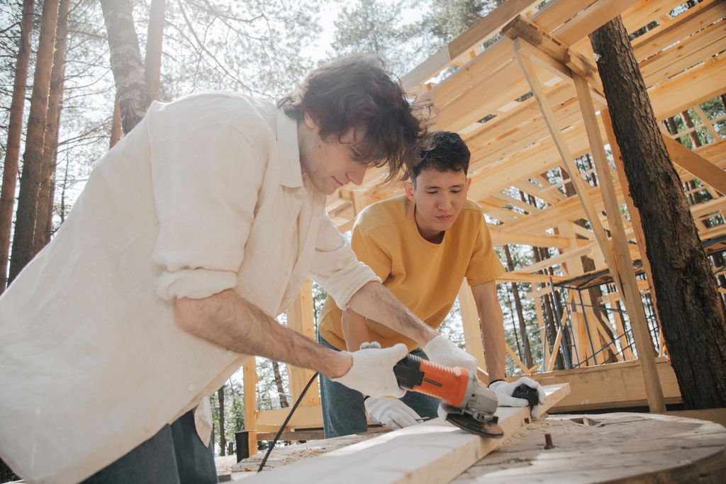 Two young builders cutting a timber board with a power tool beside the wooden frame of a house going up in a pine forest